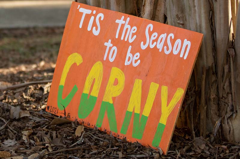 Cornhuskin Sign leaning against tree with words Tis The Season To be Corny.